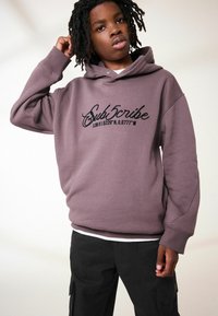 Hoodie en coton violet à coupe oversize, ornée d'un graphique "Subscribe" et de coordonnées en noir sur la poitrine, associé à un pantalon cargo noir.