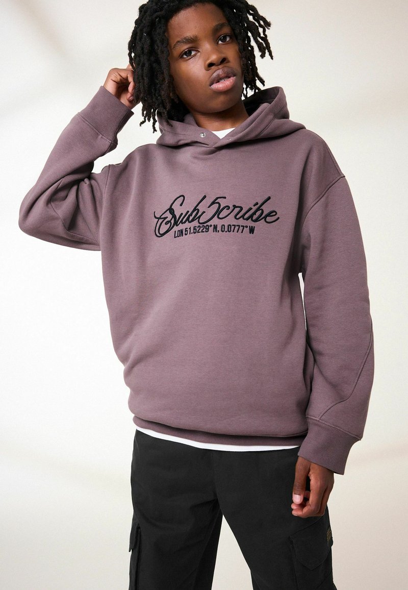 Hoodie en coton violet à coupe oversize, ornée d'un graphique "Subscribe" et de coordonnées en noir sur la poitrine, associé à un pantalon cargo noir.