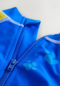 Približen posnetek modrega oblačila z zadrgo, ki ima svetlomodre odtise pasjih šap in belo šivanje blizu ovratnika.