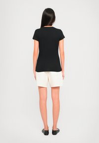 Kvinne stående med ryggen til kameraet iført en svart t-skjorte med kort erme, hvite shorts og svarte slip-on sko på en hvit bakgrunn.