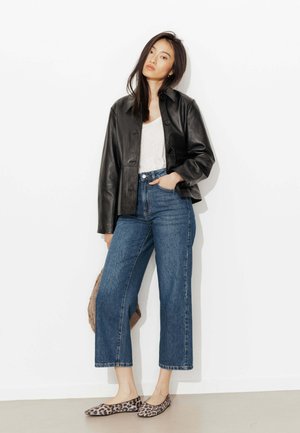 Femme portant une veste en cuir noire, un haut blanc, un jean bleu cropped et des ballerines imprimé léopard, debout contre un mur blanc, une main dans la poche.