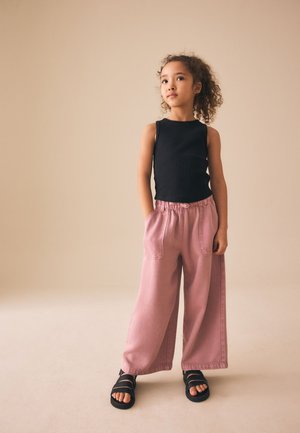 Noor tüdruk lokkis juustega, kandes musta varrukateta topi, lahtisi roosasid pükse taskutega ja musti sandaale, seisab beežil taustal.