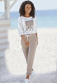 Casual white long-sleeve shirt with graphic print, beige drawstring pants, and white sneakers, set against a beach backdrop.