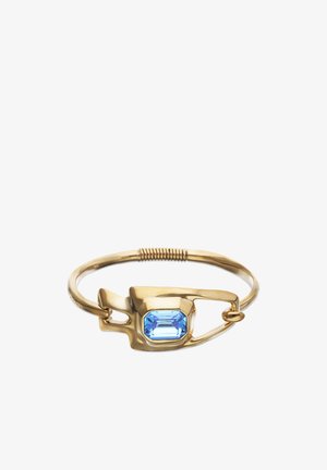 Bracelet en or conçu comme une épingle de sécurité avec une pierre précieuse bleue rectangulaire au centre, et un détail enroulé sur le bracelet.