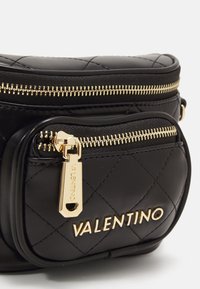 Detailní záběr na černou prošívanou koženou tašku Valentino se zlatým zipem a zlatým logem Valentino na přední kapse.