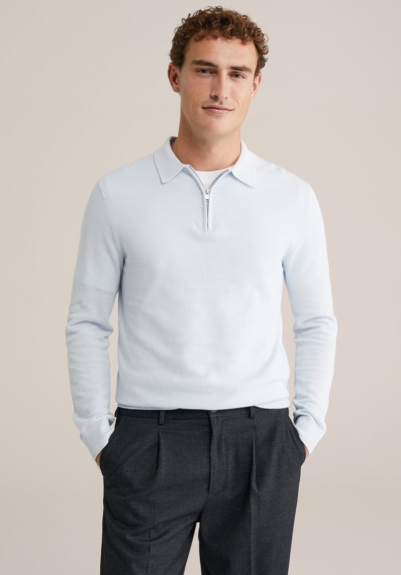 Svetlo moder pulover iz pletenine z zadrgo na izrezu, dolgimi rokavi, teksturirano tkanino, kombiniran z temno sivimi zloženkastimi hlačami, z rokami v žepih.