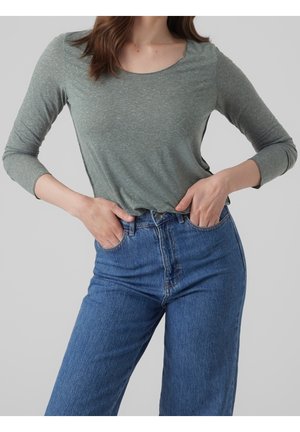 Femme portant un t-shirt gris à manches longues et un jean bleu taille haute, les mains reposant dans les poches avant sur un fond uni.
