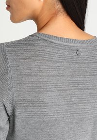 Gros plan d'une personne portant un pull gris texturé en maille avec un col rond et une petite étiquette de logo ronde en haut du dos.