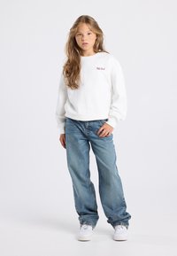 Jeune personne avec de longs cheveux portant un sweat-shirt blanc avec le texte "New York", un jean bleu ample et des baskets blanches, se tenant devant un fond uni.
