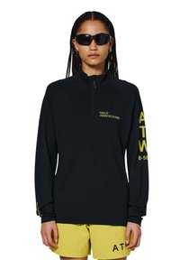 Schwarzes langärmliges Shirt mit Viertelreißverschluss und Mesh-Textur, mit gelben Textakzenten auf den Ärmeln und der Brust, kombiniert mit gelben Shorts.