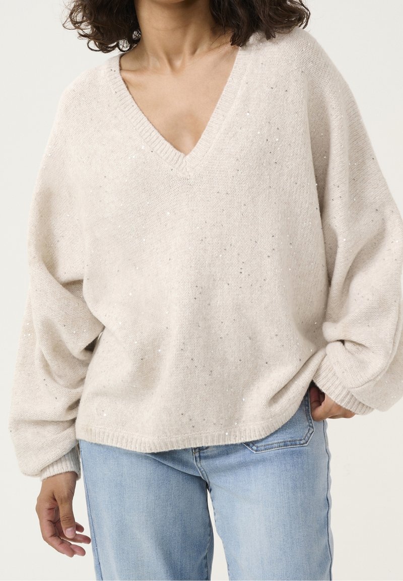 Femme portant un pull ample à col en V de couleur crème avec des paillettes subtiles et un jean bleu clair, debout avec une main dans la poche.