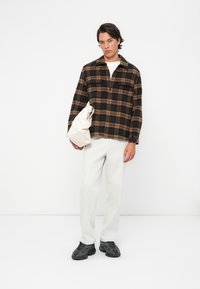 Plaid flannel shirt in black and brown, relaxed fit. Model wears white pants and holds a cream bag. Footwear is textured black shoes.