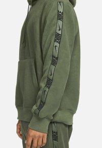 Grüne Fleece-Hoodie mit Kängurutasche, verstellbarem Kordelzug an der Kapuze und schwarzem Nike-Logo-Band entlang der Ärmel. Glatte Textur.