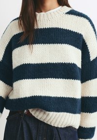 Donna che indossa un maglione spesso a maglia con ampie strisce orizzontali blu navy e crema, abbinato a jeans blu scuro e una maglietta bianca sottostante.