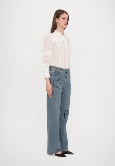 Lichte, gestructureerde, gerimpelde witte blouse gecombineerd met lichtblauwe wijde jeans met gestikte hartpatches. Zwarte puntige schoenen.