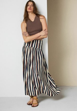 Woman wearing a sleeveless brown knit top, long skirt with vertical blue, brown, and white stripes, and tan strappy sandals standing with arms crossed.