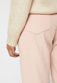 Ett hand med ljus hudton vilar på sömmen av mjuka, randiga, pastellrosa byxor, delvis skymd av en chunky krämfärgad tröja.