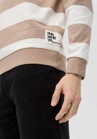 Beżowa i biała bluza w paski z bawełny, z prążkowanymi mankietami oraz metką marki po dolnej stronie. Zestawiona z czarnymi spodniami.