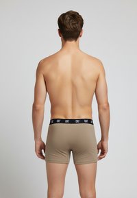 Beige boxer shorts with a black elastic waistband displaying "CR7," viewed from the back on a male model. Smooth fabric texture.