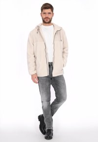 Chaqueta de forro polar beige con cremallera frontal, dos bolsillos laterales y un panel contrastante en el pecho superior. Se lleva con vaqueros grises y botas negras.