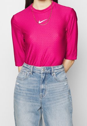 Frau trägt ein leuchtend pinkes, strukturiertes Nike-Shirt mit ellbogenlangen Ärmeln, goldener Kettenhalskette und hellblauen, hoch taillierten Jeans.