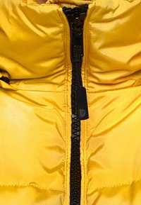 Gele pufferjas met een glanzende, doorgestikte structuur. Heeft een centrale zwarte ritssluiting en een hoge kraag voor extra warmte.