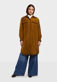 Hnedá košela s dlhým rukávom vo forme bundy vyrobená z hladkého materiálu, s dvoma náprsnými vreckami a oblým lemom, skombinovaná s voľnými modrými rifľovými nohavicami.