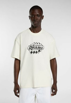 Hombre vestido con camiseta blanca extragrande de Dickies y pantalones blancos, de pie frente a un fondo liso gris claro.