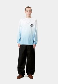 Camisa de manga larga con un degradado azul que va del blanco al azul claro, con un logo negro en el pecho, combinada con pantalones anchos negros y zapatos marrones.