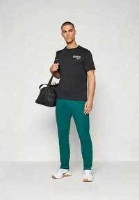 Mann in schwarzem Reebok-T-Shirt, türkisfarbigen Jogginghosen, weißen Sneakers, der eine schwarze Sporttasche über der Schulter trägt und vor einem einfarbigen hellen Hintergrund steht.