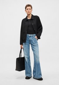 Casaco de mistura de lã preta com gola e botões, combinado com jeans azul claro com pernas evasê. Modelo segura uma mala tote de couro preta.