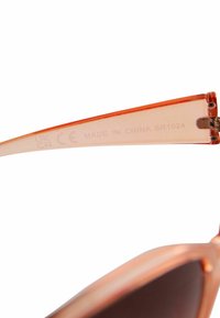 Un bras de lunettes de soleil orange transparent avec des accents métalliques, comportant le texte imprimé "FABRIQUÉ EN CHINE SR1024" sur le côté intérieur.