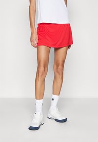Rode sportrok met een gladde textuur, gecombineerd met een wit topje en witte sneakers met blauwe accenten en geribde witte sokken.
