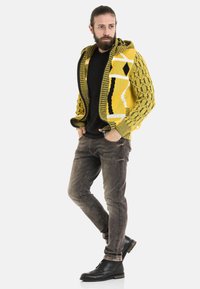 Gelbe gemusterte Strickjacke mit einem strukturierten Diamantdesign, kombiniert mit dunklen Jeans und schwarzen Schnürstiefeln.