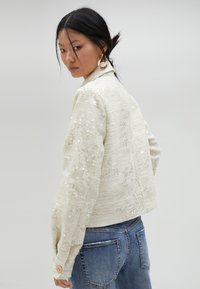 Kort, krämfärgad jacka av texturerad tyg med paljetter. Har en hög krage, långa ärmar och knäppta manschetter.