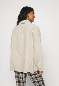 Chaqueta de camisa de pana beige con un ajuste relajado, gran cuello, detalles de costura visibles y puño abotonado, combinada con pantalones de cuadros en blanco y negro.