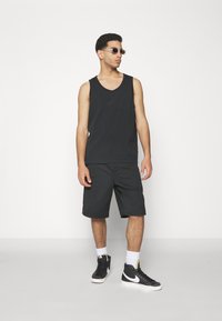 Melnas bez piedurknēm tank top ar smalku logotipu, kombinēts ar melniem šortiem līdz ceļiem. Valkā ar melniem augstiem apaviem un caurspīdīgām saulessargiem.