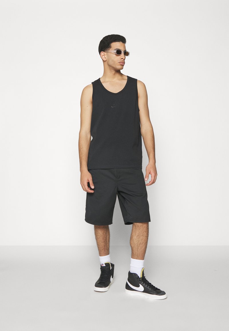 Camiseta sin mangas negra con un logo sutil, combinada con pantalones cortos negros hasta la rodilla. La prenda se lleva con zapatillas altas negras y gafas de sol transparentes.