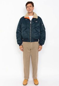 Marineblaue Bomberjacke mit einem Schafsfellkragen, orangefarbenem Innenfutter, Reißverschluss vorne und zwei übergroßen Taschen; getragen mit hellbeigen Hosen und beigefarbenen Schuhen.