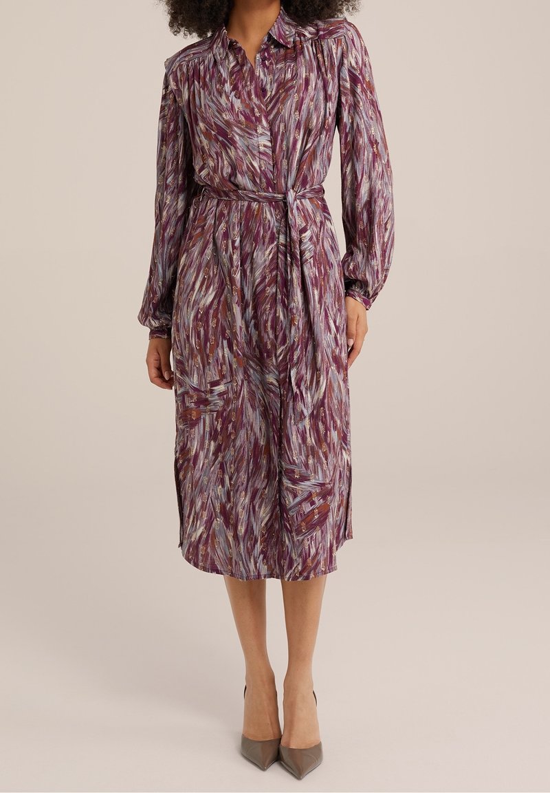 Purple and multicolored patterned dress with long sleeves, button-down front, waist tie, and side slits, paired with pointed-toe heels.