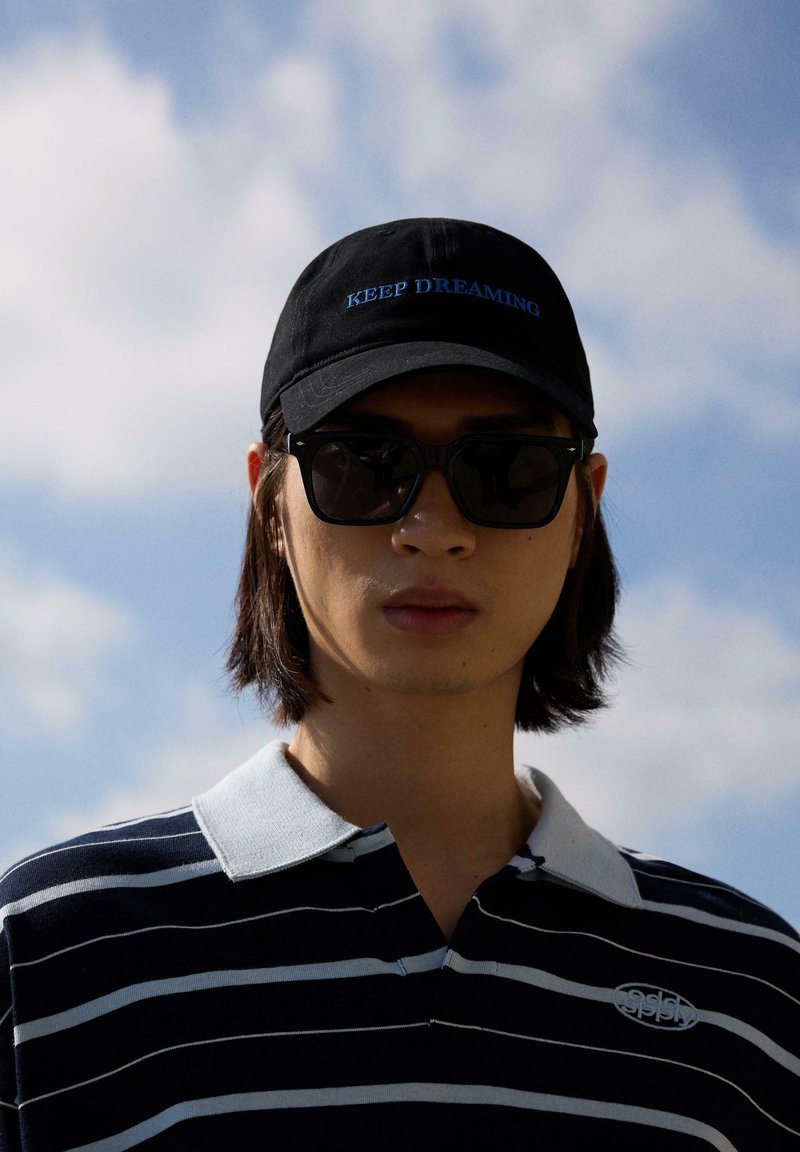 Jeune personne portant une casquette noire avec le texte "KEEP DREAMING", des lunettes de soleil noires, et un polo rayé noir et blanc sur fond de ciel nuageux.