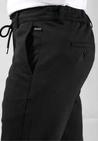 Zwarte joggingsbroek met elastische tailleband en trekkoord. Voorzien van slanke zijzakken en een klein logo-label aan de voorkant. Glad textuur.