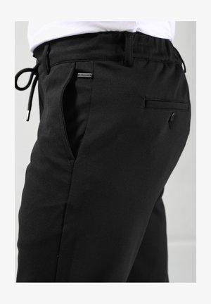 Zwarte joggingsbroek met elastische tailleband en trekkoord. Voorzien van slanke zijzakken en een klein logo-label aan de voorkant. Glad textuur.