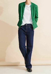 Grüne Lederjacke mit gerippten Schultern, weißem T-Shirt und dunkelblauen geraden Jeans. Schwarze Schuhe ergänzen das Outfit.