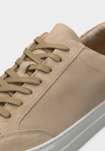 Μπεζ σουέντ αθλητικό παπούτσι με κορδόνια στο ίδιο χρώμα και λευκή λαστιχένια σόλα, με κοντινή λήψη των ραφών και της γλώσσας.