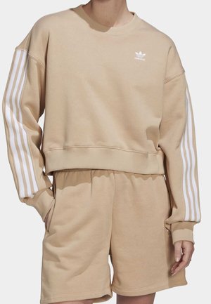 Beżowa bluza Adidas z białymi paskami na rękawach i dopasowanymi beżowymi szortami, noszona przez osobę z rękami w kieszeniach.