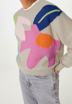 Person iført beige striktrøje med store farverige abstrakte blomsterprint kombineret med lysegrå jeans, hånd i lommen.