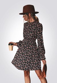 Zwarte bloemenjurk met lange mouwen en een gathered taille, met een roze en groen bloemenpatroon. Een bruine hoed en beker maken de outfit compleet.