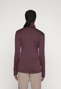 Persona con capelli intrecciati legati indietro, che indossa una maglietta attillata a maniche lunghe color bordeaux e pantaloni beige, girata di spalle contro uno sfondo semplice.