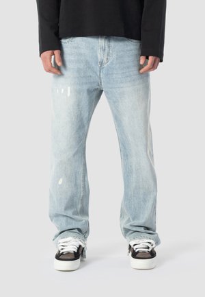 Hellblaue, locker sitzende Jeans mit dezenten weißen Farbspritzern, kombiniert mit schwarz-weißen Turnschuhen und einem schwarzen Langarmshirt.